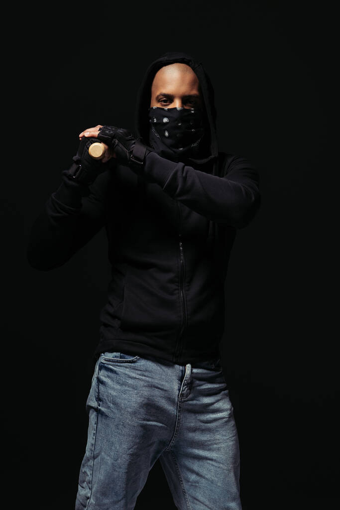 Africano americano hooligan em luvas e máscara facial segurando bastão de beisebol isolado em preto  - Foto, Imagem