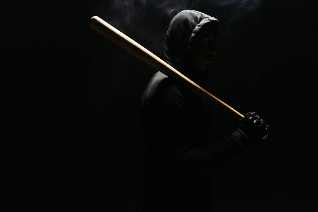 Silhouette of bandit in hoodie holding baseball bat on black background with lighting and smoke  - 写真・画像