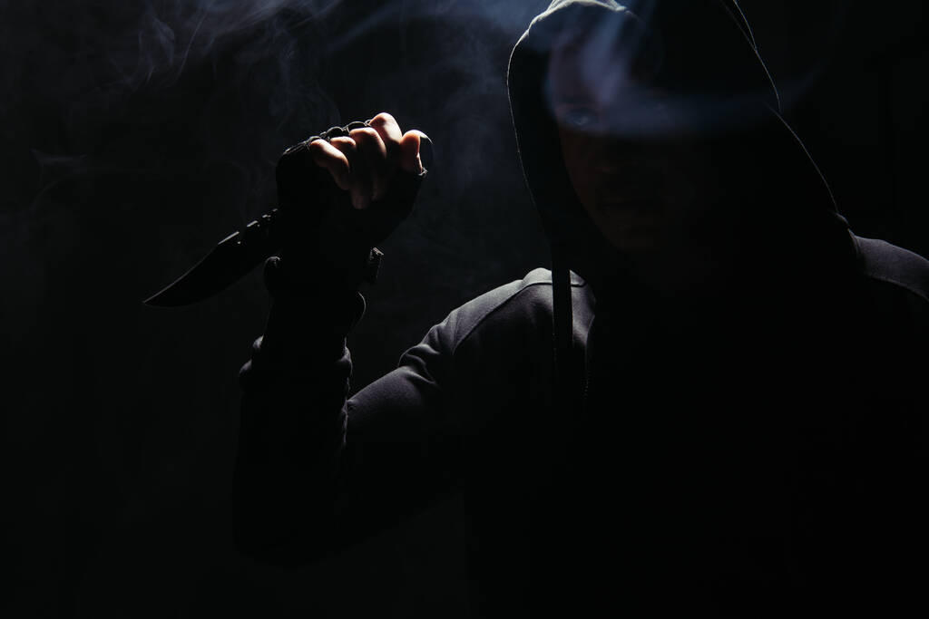 Silhouette of african american bandit in hood holding knife on black background with smoke  - Foto, imagen