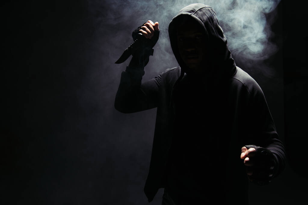 Silhouette of african american bandit in hoodie holding knife on black background with smoke  - Fotó, kép