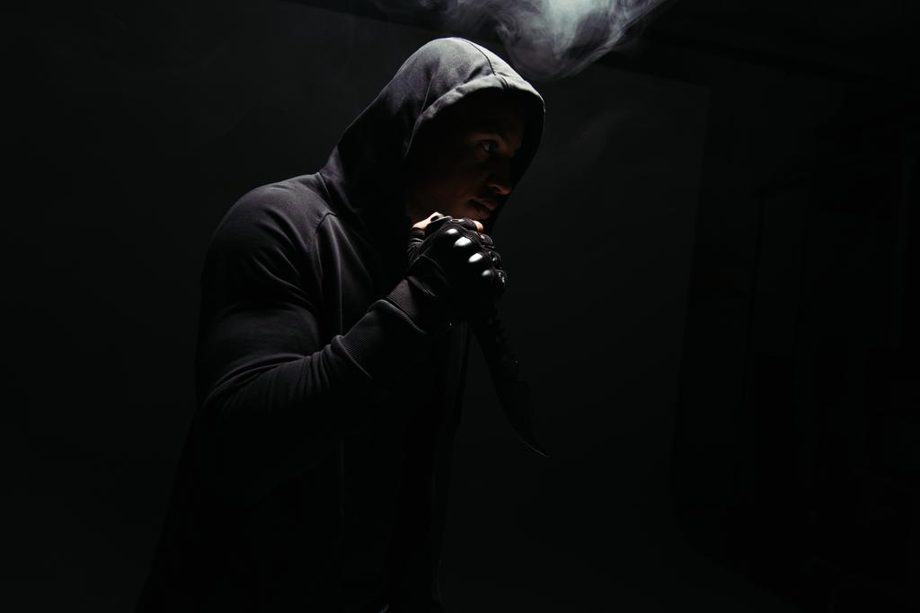 Silhouette of african american bandit holding knife on black background with smoke  - 写真・画像