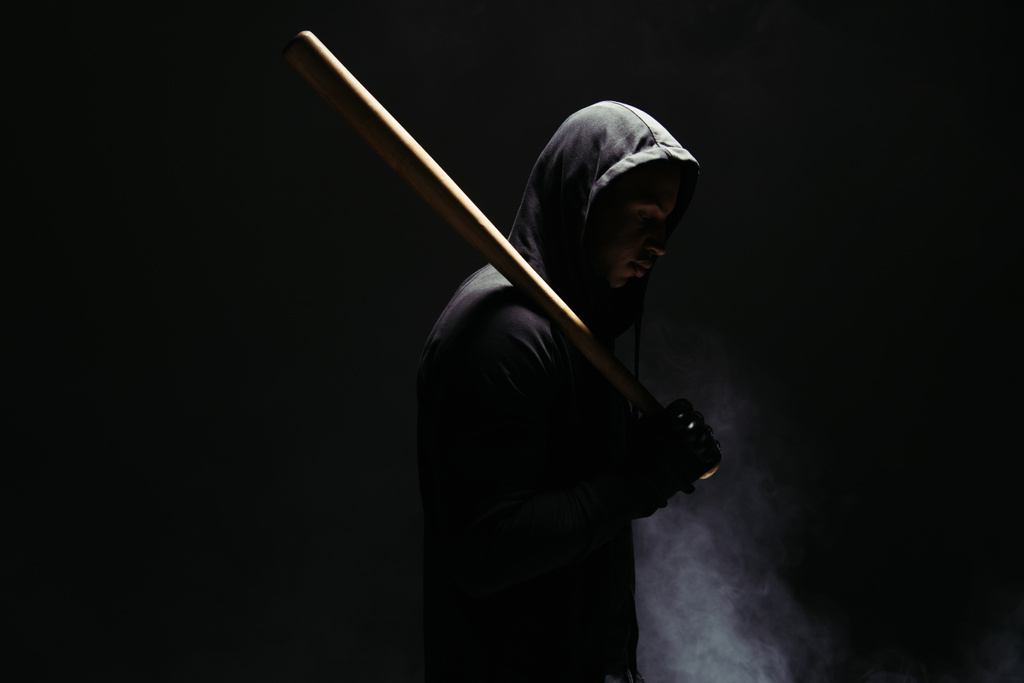 Вид сбоку на африканского американского бандита в капюшоне с деревянной бейсбольной битой на черном фоне с дымом  - Фото, изображение