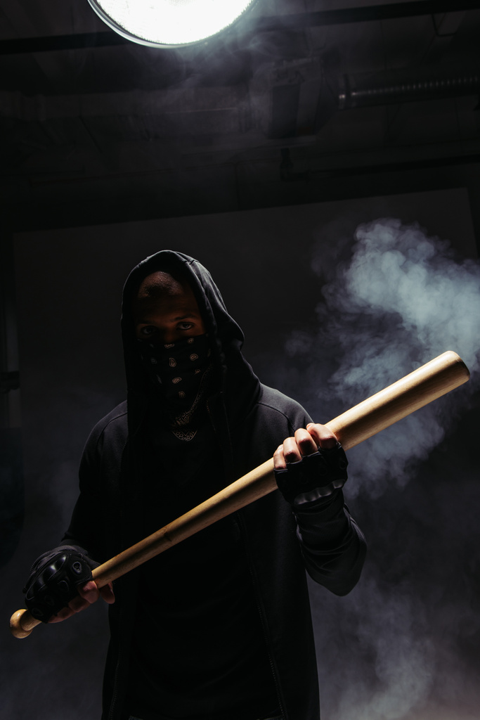 Silhouette of african american vandal with mask on face holding baseball bat on black background with smoke  - Foto, Imagem