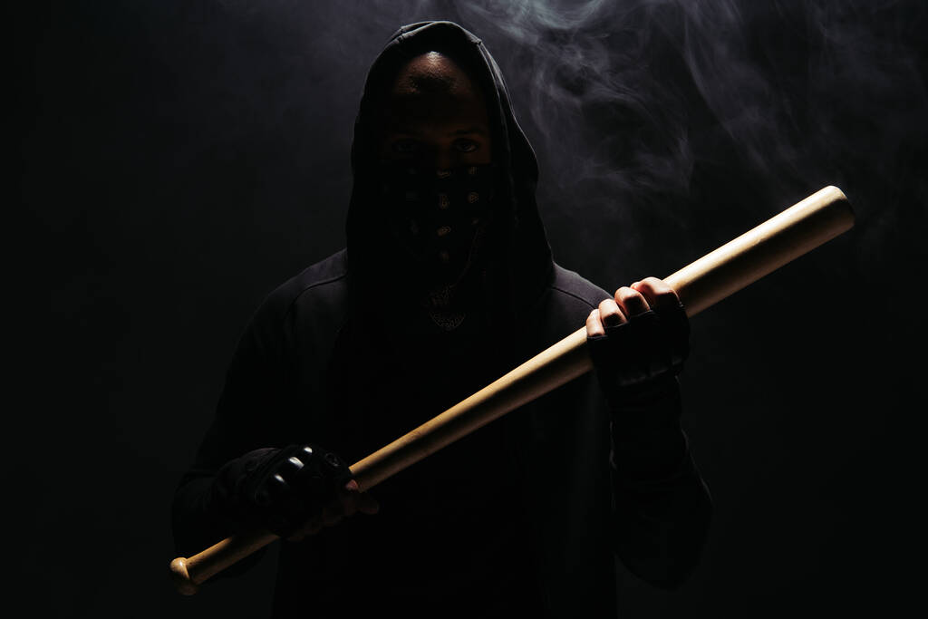 Silhouette of african american man in hoodie and gloves holding baseball bat on black background with smoke  - Valokuva, kuva