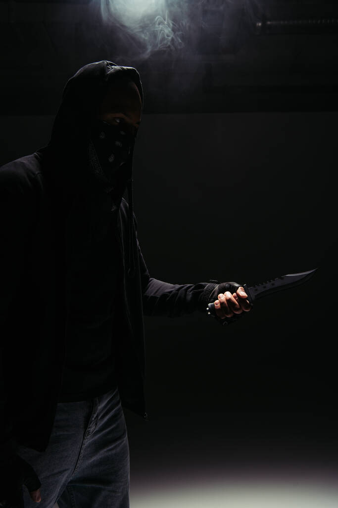 Silhouette di rapinatore afroamericano in maschera e felpa con cappuccio con coltello su sfondo nero con fumo  - Foto, immagini