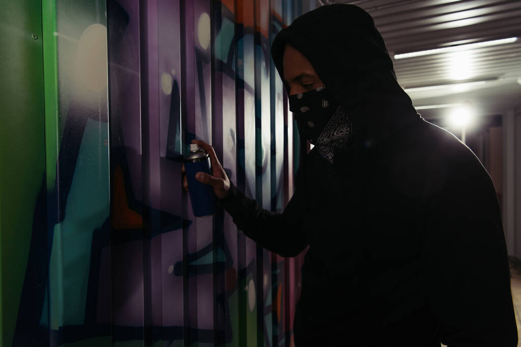 Side view of african american hooligan in hoodie and mask drawing graffiti on wall  - Photo, Image