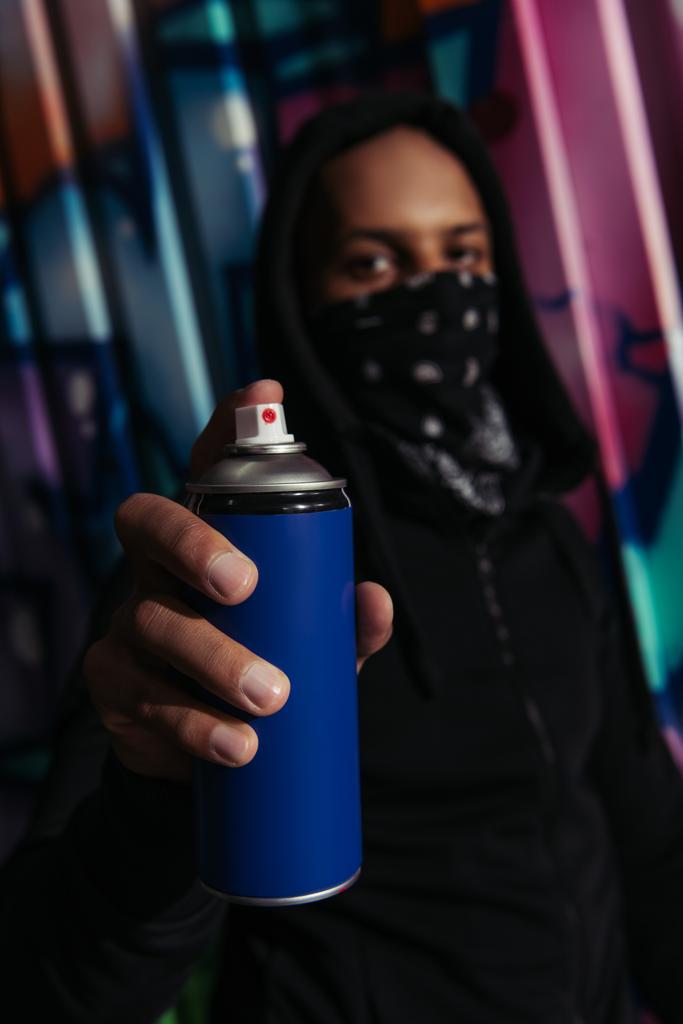Размытый африканский американский вандал с маской на лице, держащий распылитель краски рядом с граффити на городской улице  - Фото, изображение