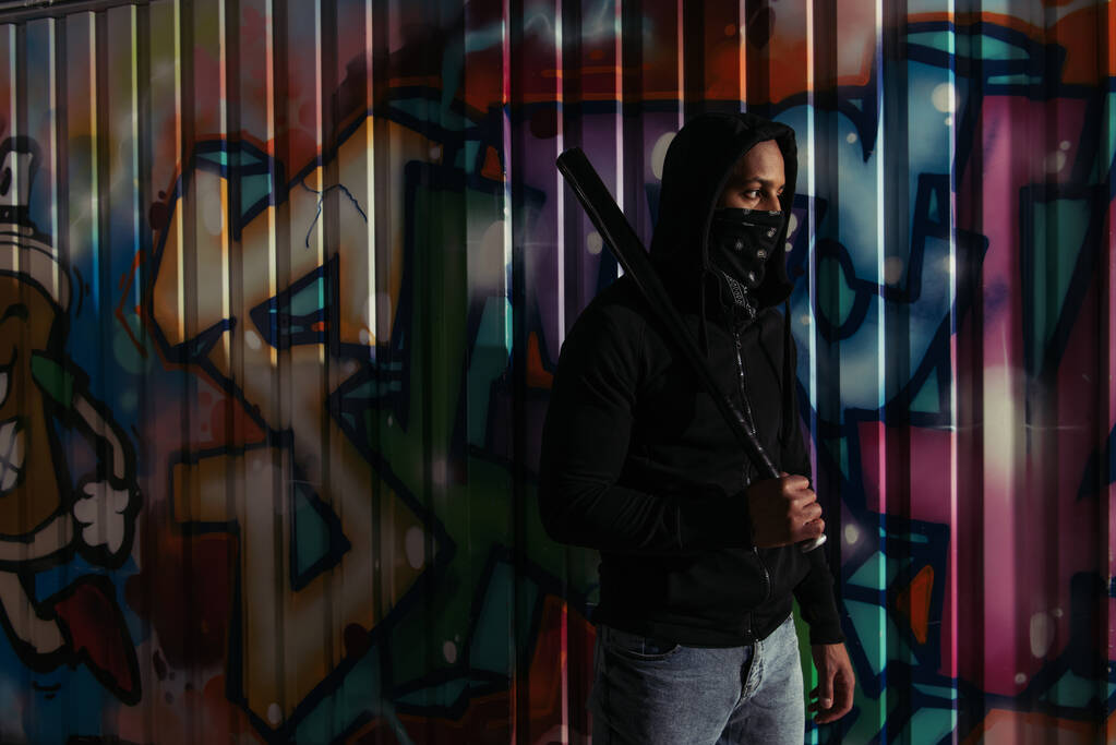 Afrikai amerikai vandál maszkkal az arcán baseball ütőt tart a graffiti közelében a városi utcán  - Fotó, kép