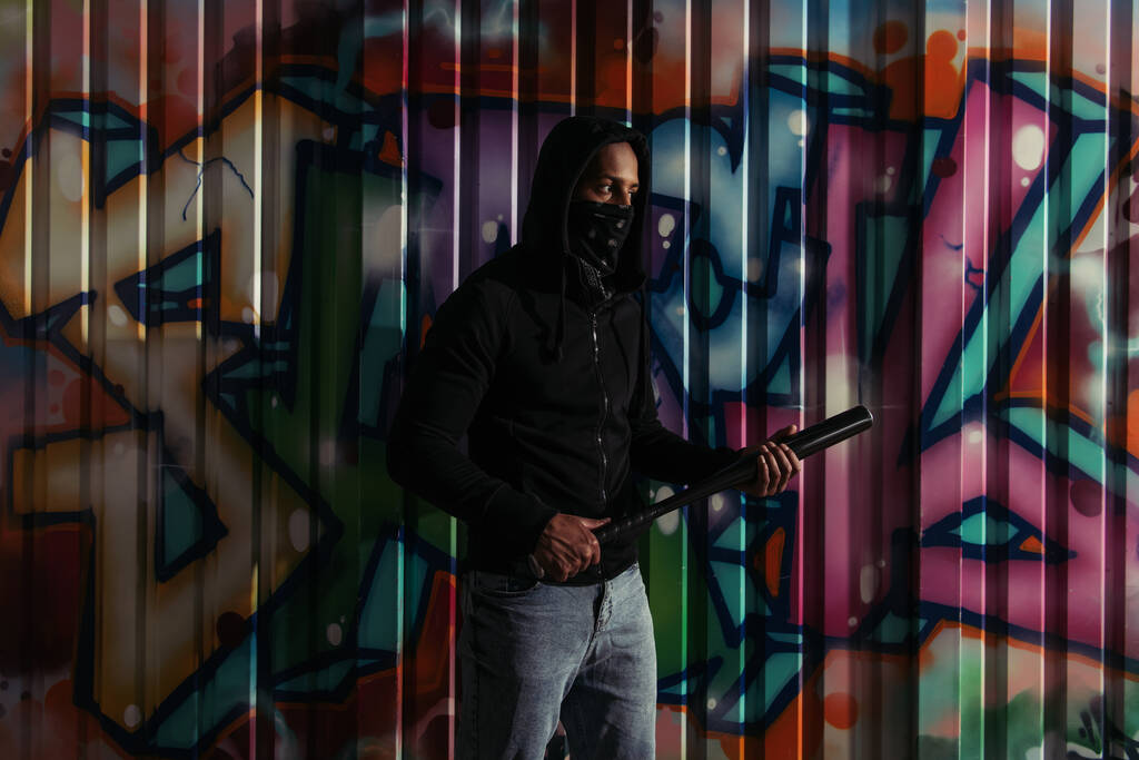 Африканський американець - вандал у худі, що тримає бейсбольну биту біля графіті вночі.  - Фото, зображення