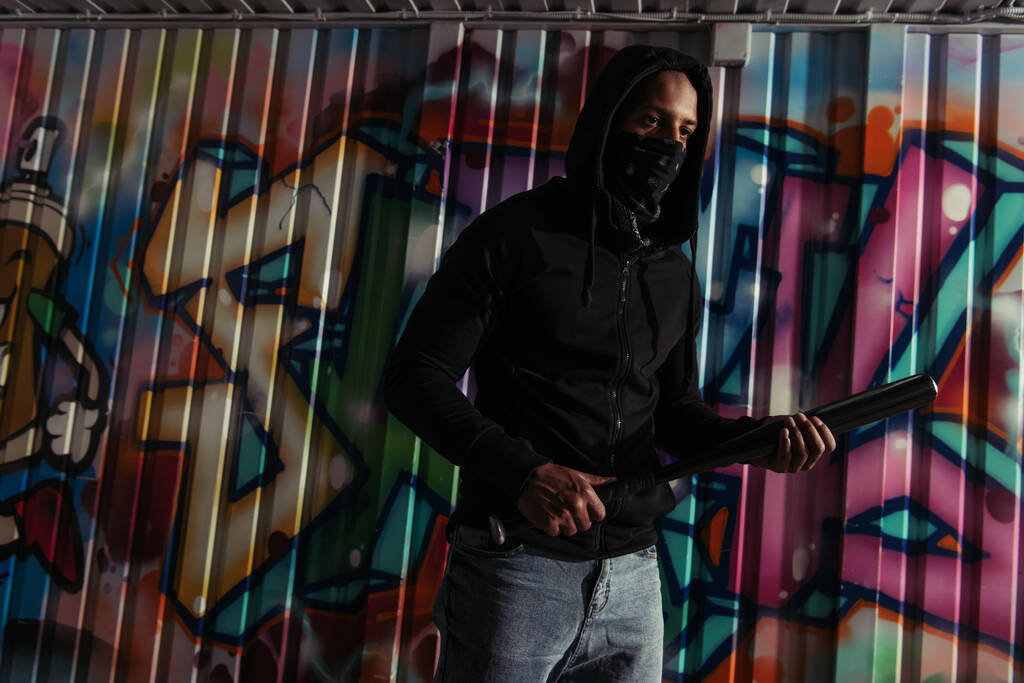Gamberro afroamericano con capucha sosteniendo bate de béisbol cerca de graffiti en la pared  - Foto, imagen