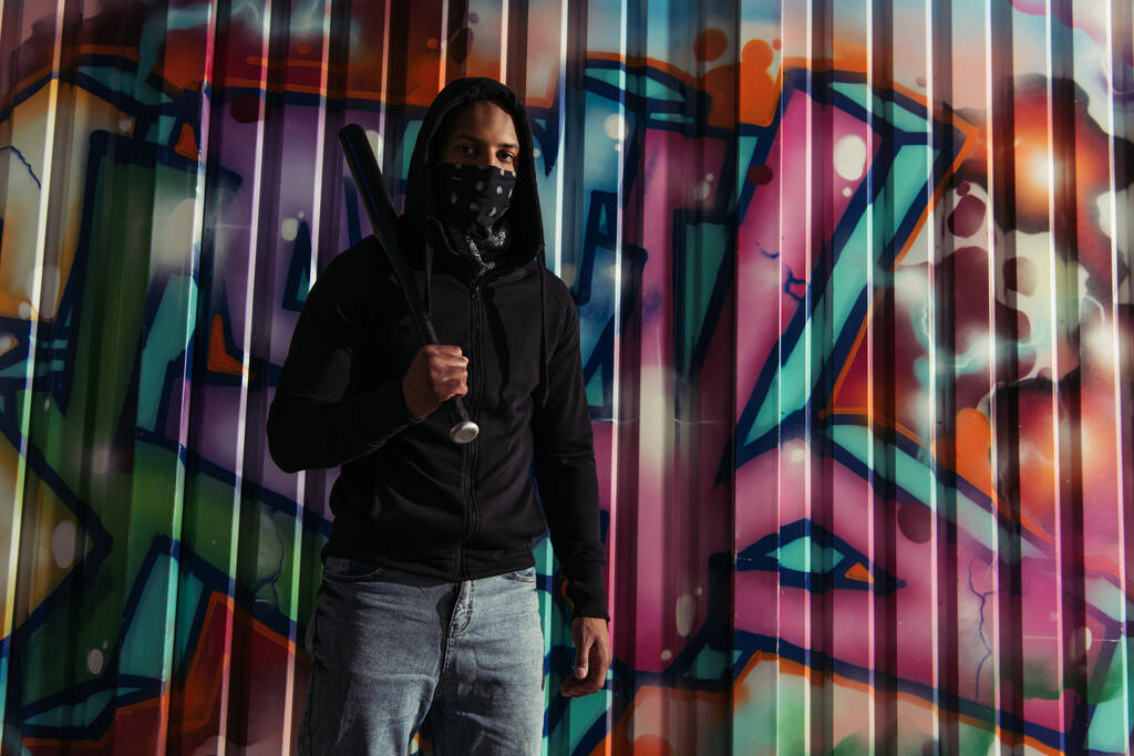 African american hooligan holding baseball bat and looking at camera near graffiti outdoors  - Zdjęcie, obraz