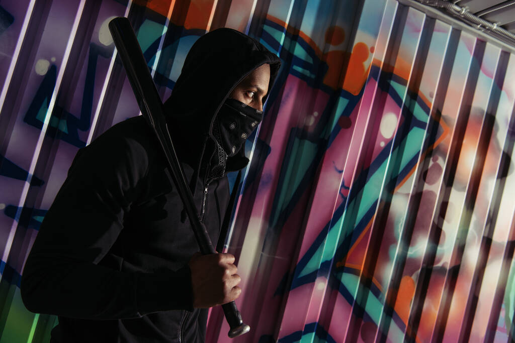 Side view of african american bandit in mask holding baseball bat near graffiti on wall  - 写真・画像