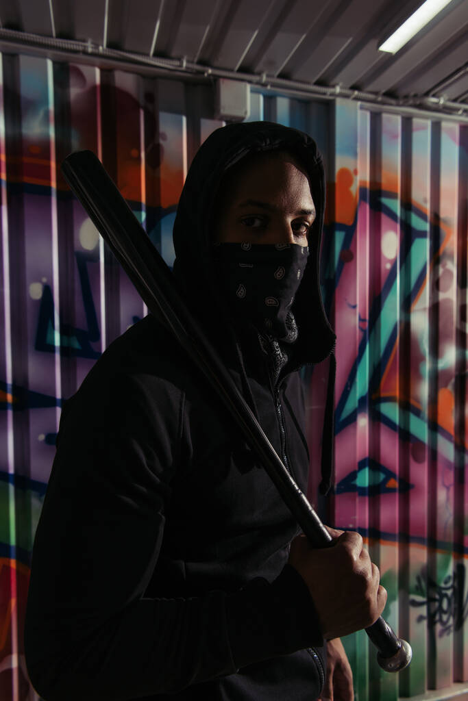 Afrikanischer Hooligan mit Baseballschläger schaut in die Kamera neben Graffiti an Wand  - Foto, Bild