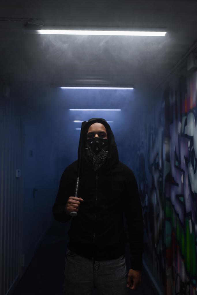 African american hooligan in mask holding baseball bat and looking at camera near lighting and graffiti  - Fotoğraf, Görsel