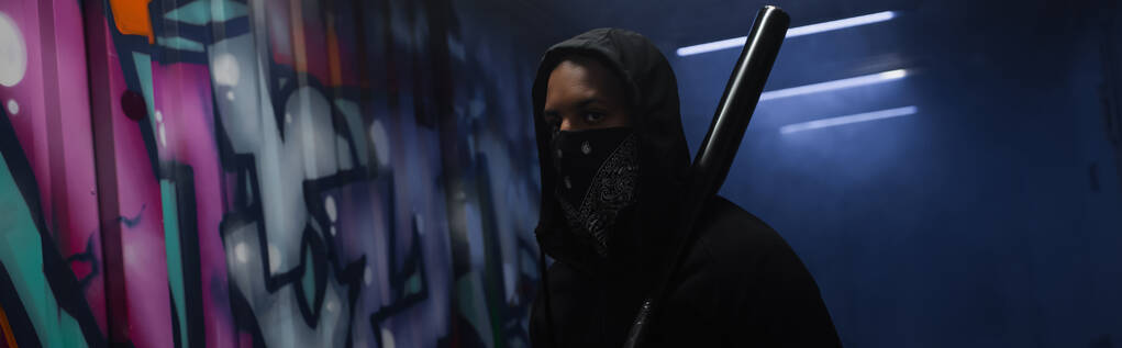African american bandit with mask on face holding baseball bat near graffiti in garage with smoke, banner  - Valokuva, kuva