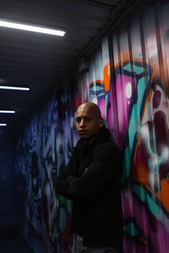 Gamberro afroamericano cruzando brazos cerca de graffiti en pared en garaje  - Foto, imagen