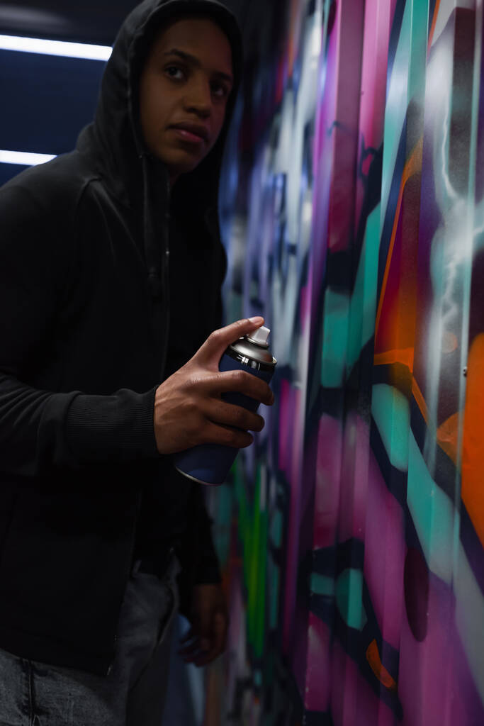 Blurred african american vandal in hood painting graffiti and looking away in garage  - 写真・画像