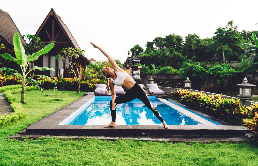 Donna in abbigliamento sportivo che fa yoga piegandosi lateralmente mentre si esercita vicino alla piscina in giardino con alberi esotici e cespugli sul resort - Foto, immagini