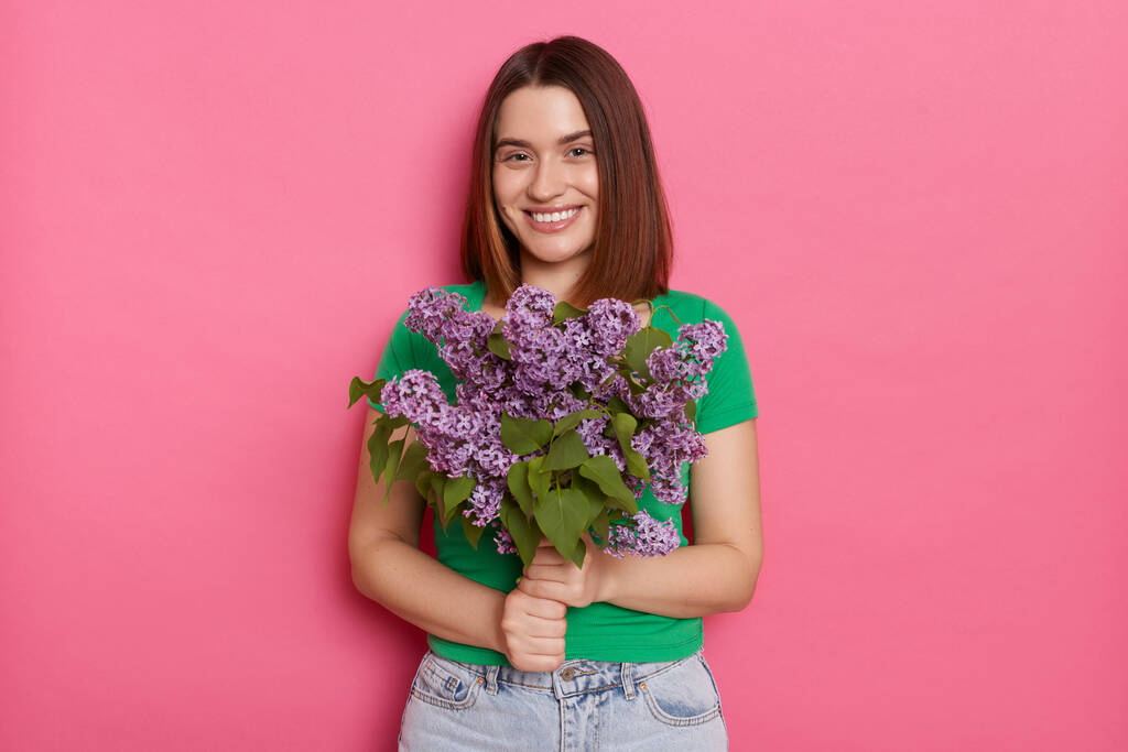 Photo of pleased dark haired woman wearing green T-shirt standing looking with pleasure at camera, carries lilac flowers, models against pink background. Spring present. - Fotografie, Obrázek