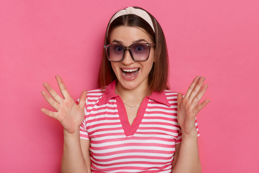 Horizontal shot of excited surprised woman wearing striped T-shirt and sunglasses posing isolated over pink background, standing with raised arms, being pleasantly surprised. - Photo, Image