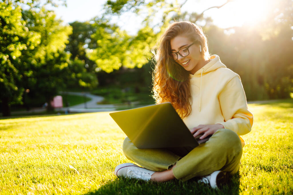 Jeune femme avec casque sans fil travaillant en ligne ou étudiant assis sur l'herbe au parc. Entreprise, blogging, freelance, concept d'éducation. - Photo, image