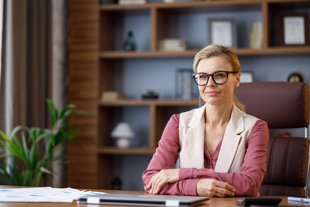 Portrait of beautiful blondie woman looking at camera and smiling. Adult stylish confident businesswoman wearing eyeglasses sitting at workplace in office. Portrait of modern successful female leader. - Foto, Imagen