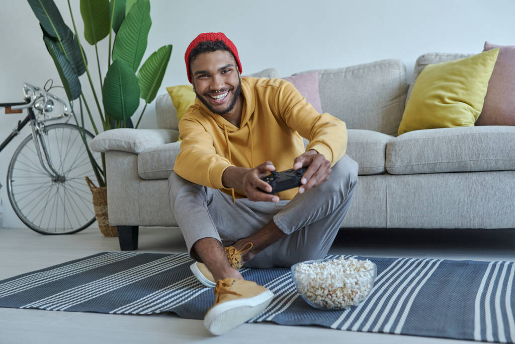 Joyful young African man playing video games while sitting on the floor at home - Zdjęcie, obraz