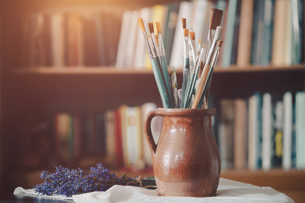 Still life of paint brushes  in clay jug, old books in the background. - Photo, image