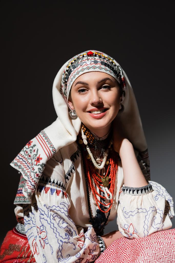 portret van gelukkige jonge Oekraïense vrouw in traditionele kleding met ornament en rode kralen op zwart  - Foto, afbeelding
