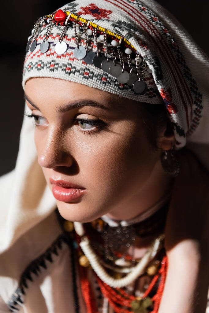 portrait of young ukrainian woman in traditional headwear with ornament and red beads on black  - Fotografie, Obrázek