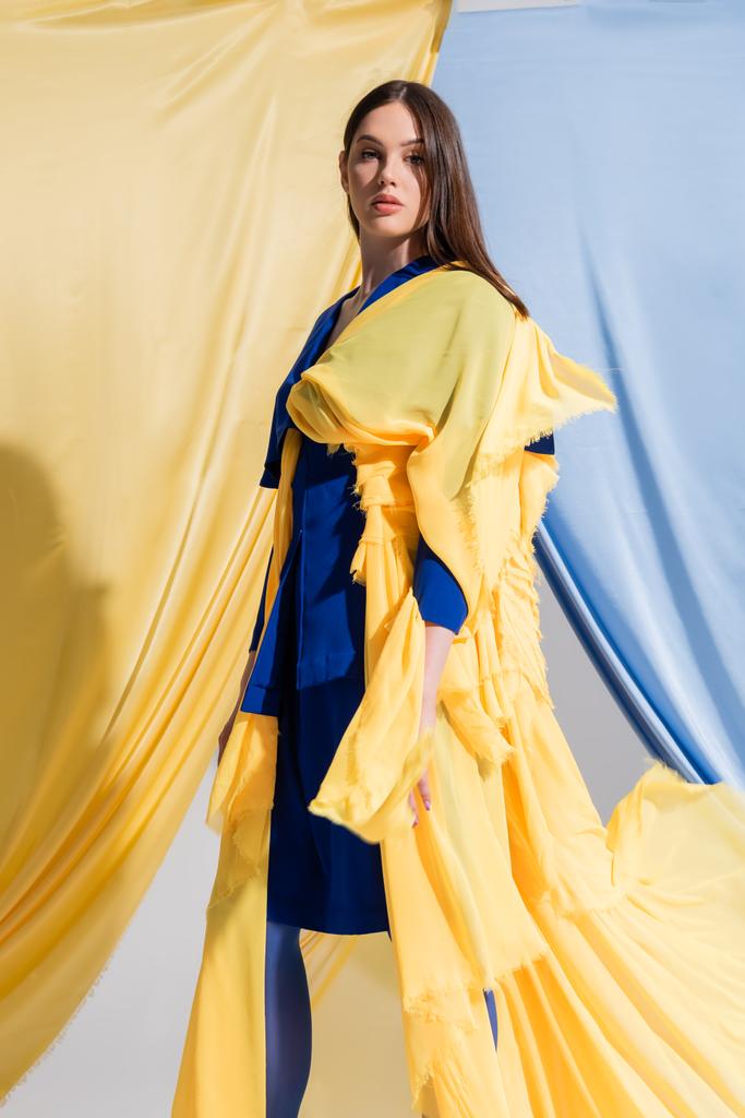 junge Ukrainerin in farbigem Blockkleid posiert in der Nähe blauer und gelber Vorhänge  - Foto, Bild
