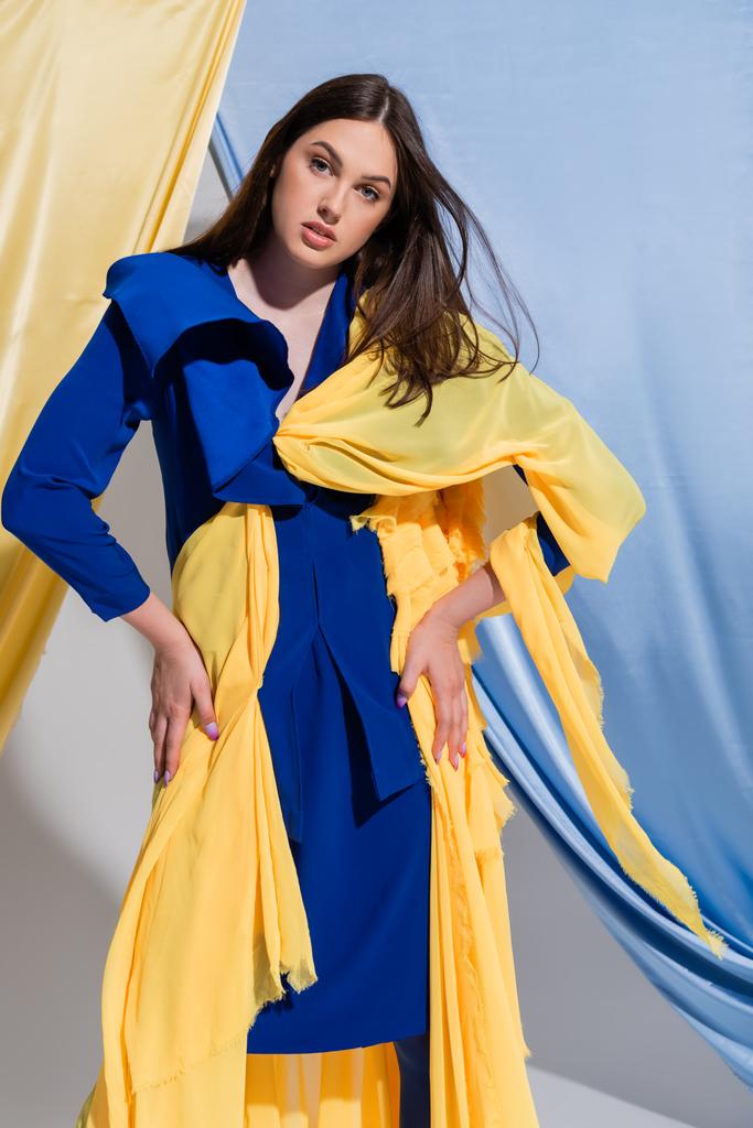 young ukrainian woman in color block dress posing with hands on hips near blue and yellow curtains  - 写真・画像