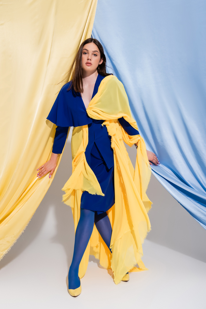 Повна довжина стильної українки в кольоровому сукні, що поставляється біля синіх і жовтих завіс.  - Фото, зображення