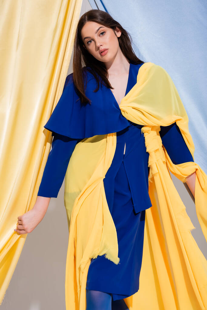 trendige junge Ukrainerin in farbigem Blockkleid posiert in der Nähe blauer und gelber Vorhänge  - Foto, Bild
