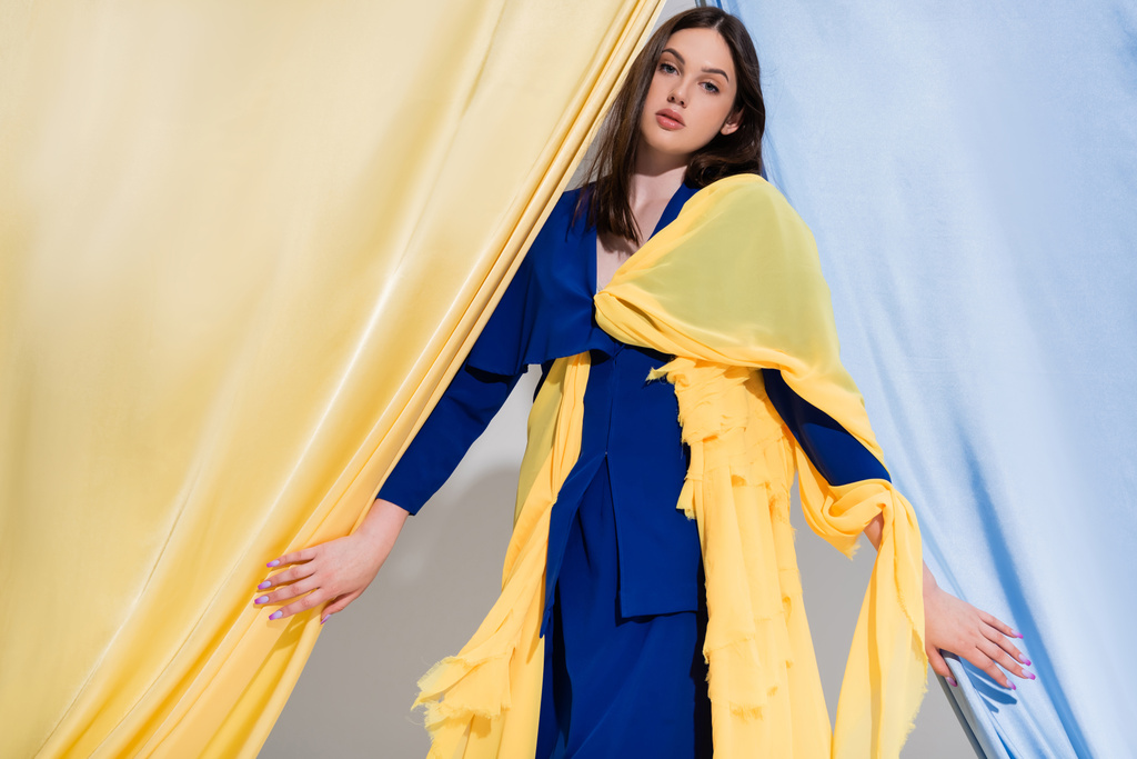 jovem mulher ucraniana elegante em vestido de bloco de cor posando perto de cortinas azuis e amarelas  - Foto, Imagem