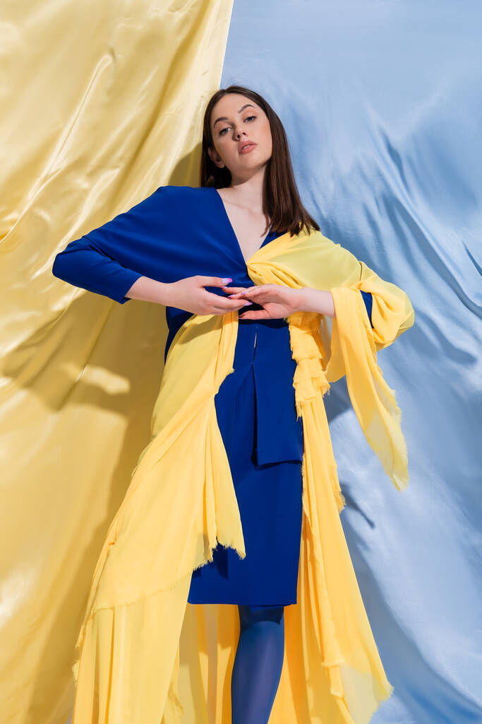 mujer ucraniana joven y confiada en ropa de bloque de color posando cerca de cortinas azules y amarillas  - Foto, imagen