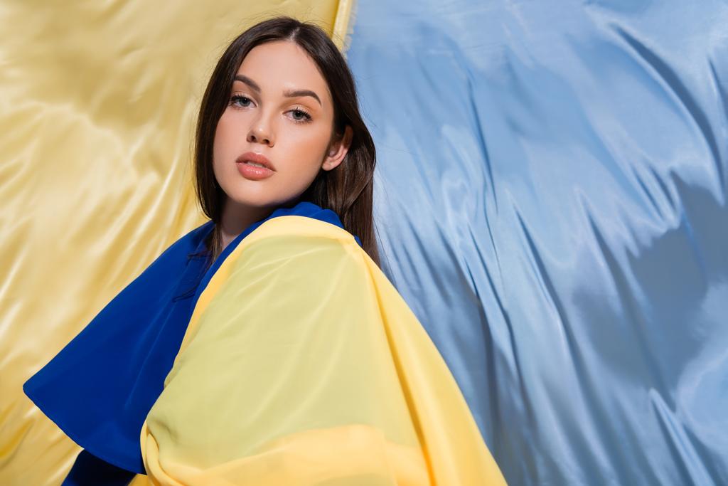 πατριωτική νεαρή Ουκρανή γυναίκα με ρούχα μπλοκ χρώματος που ποζάρουν κοντά σε μπλε και κίτρινη σημαία - Φωτογραφία, εικόνα
