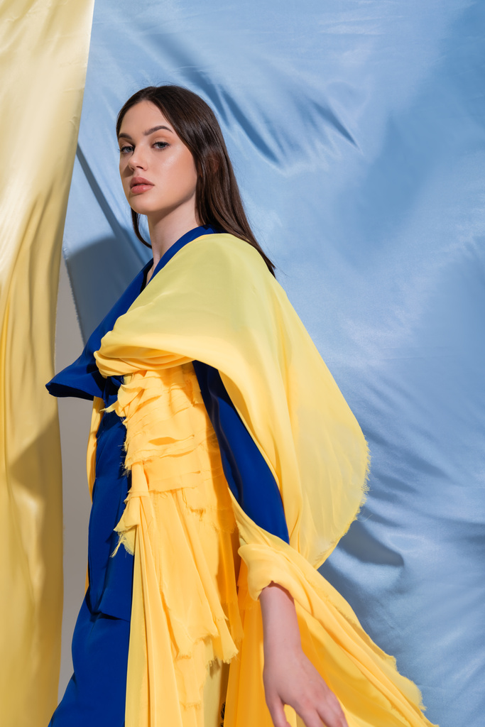 молодая украинка в цветной блочной одежде стоит рядом с сине-желтой тканью - Фото, изображение