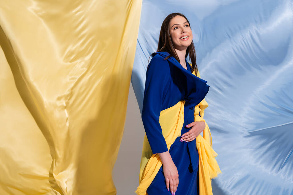 joyful young ukrainian woman in color block clothing posing near blue and yellow fabric - Foto, afbeelding