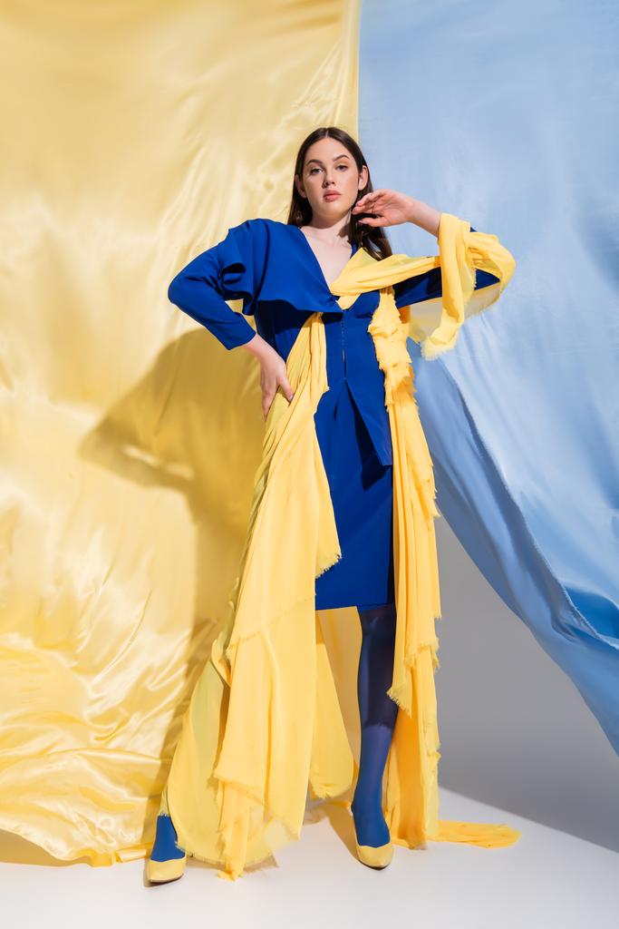 πλήρες μήκος της ουκρανικής γυναίκας στη μόδα χρώμα μπλοκ ένδυσης ποζάρουν με το χέρι στο ισχίο κοντά στο μπλε και κίτρινο ύφασμα - Φωτογραφία, εικόνα
