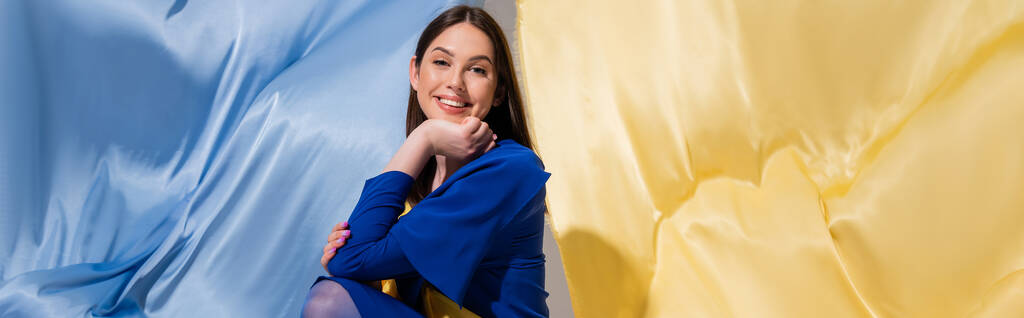 positieve Oekraïense vrouw in stijlvolle kleur blok kleding poseren in de buurt van blauwe en gele vlag, banner - Foto, afbeelding