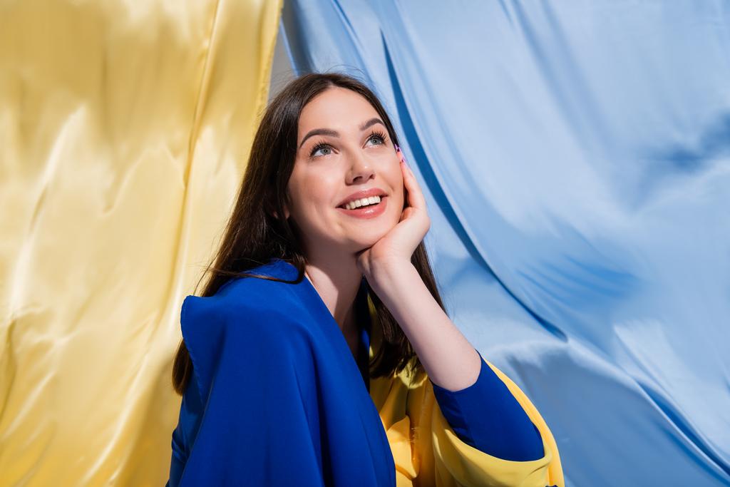 dreamy ukrainian woman in stylish color block clothing smiling near blue and yellow flag - Foto, Imagen