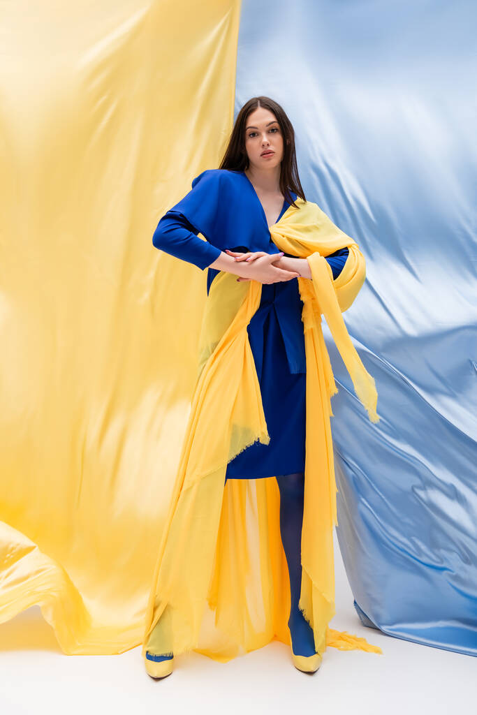 volledige lengte van Oekraïense jonge vrouw in stijlvolle kleur blok kleding poseren in de buurt van blauw en geel weefsel - Foto, afbeelding