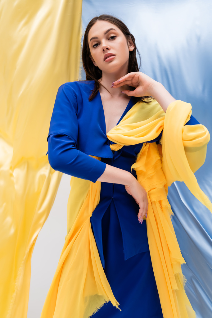 Oekraïense jonge vrouw in stijlvolle kleur blok kleding poseren in de buurt van blauw en geel weefsel - Foto, afbeelding