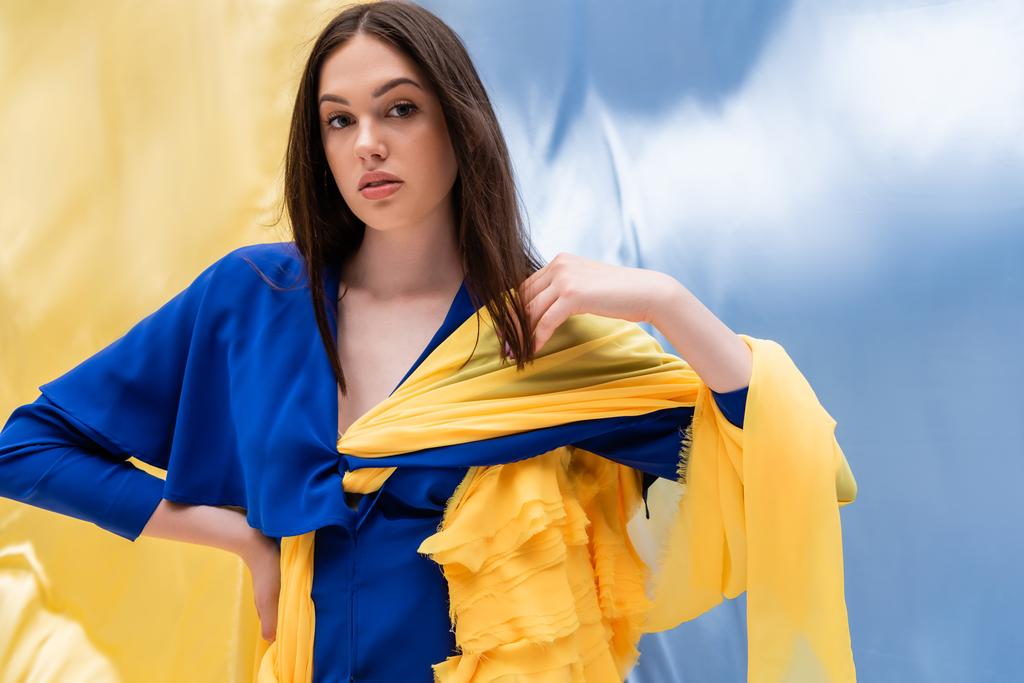 brunette young woman in stylish color block clothing posing with hand on hip near ukrainian flag - 写真・画像