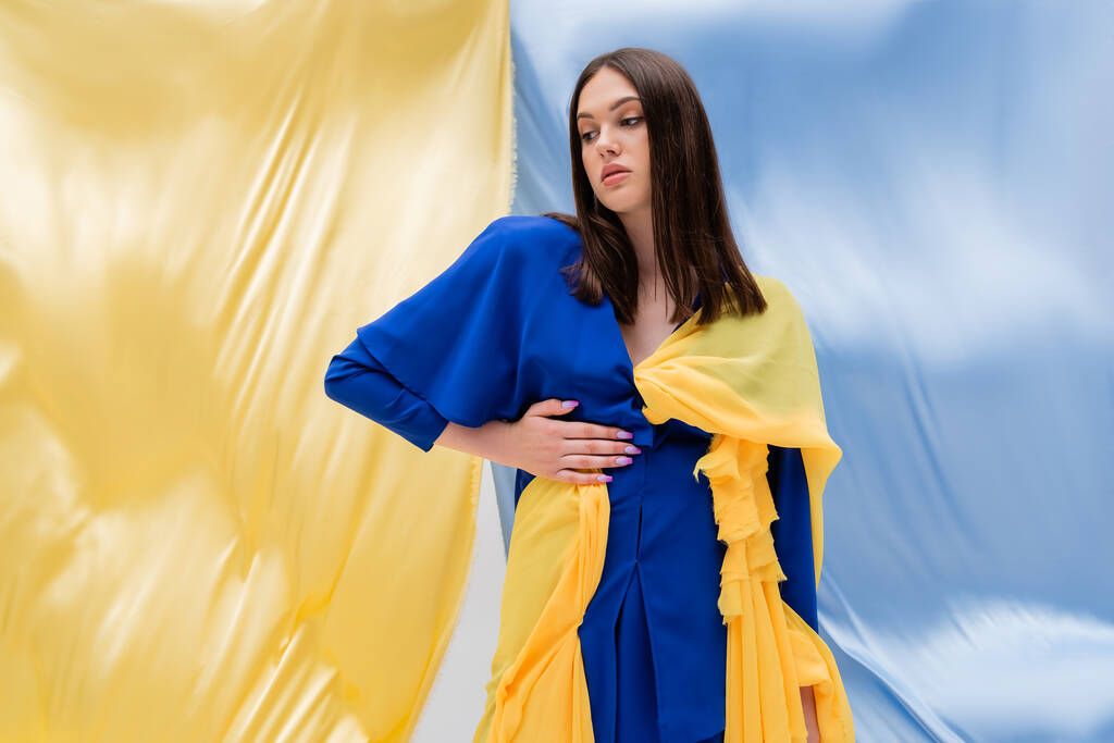pretty ukrainian young woman in stylish color block clothing standing with hand on hip near blue and yellow fabric - 写真・画像