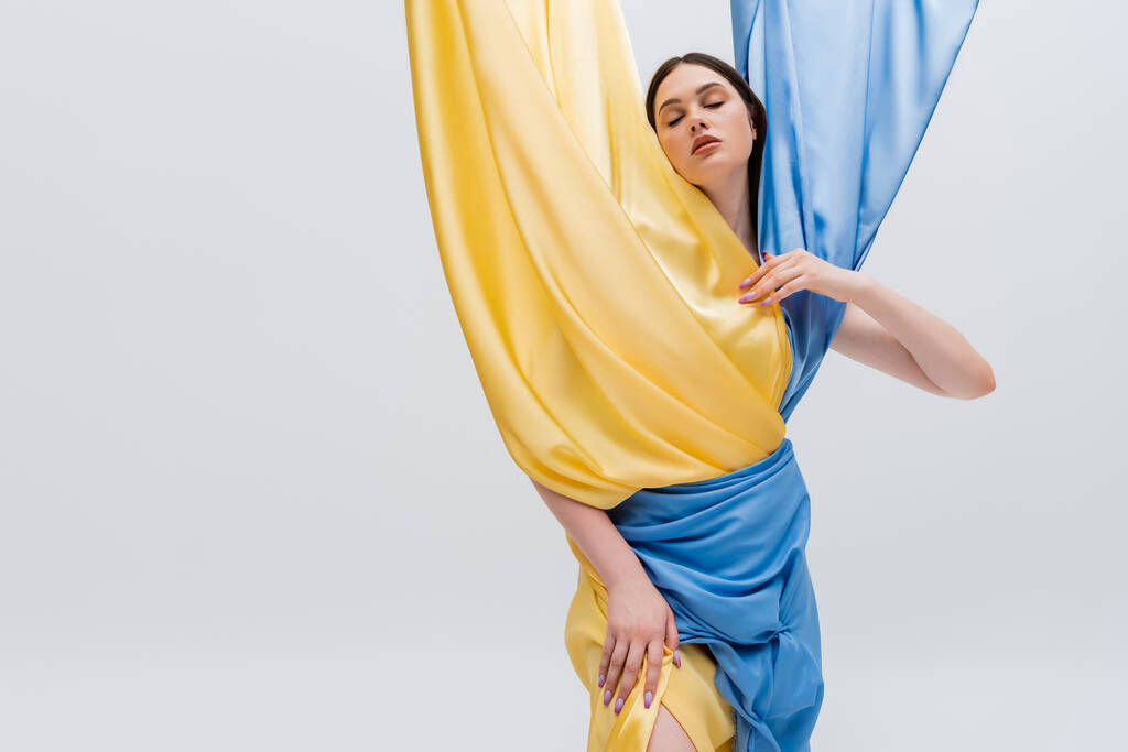 joven morena en vestido azul y amarillo posando con los ojos cerrados aislados en gris - Foto, Imagen