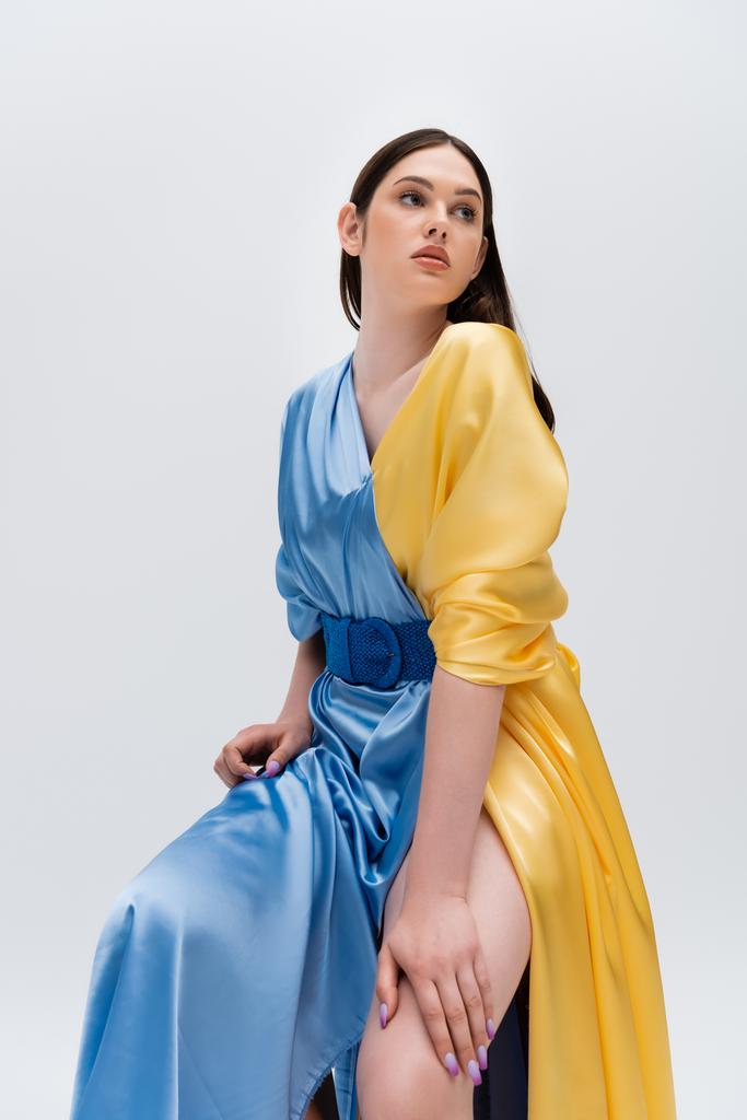 confident young ukrainian woman in blue and yellow dress posing while sitting isolated on grey - Zdjęcie, obraz
