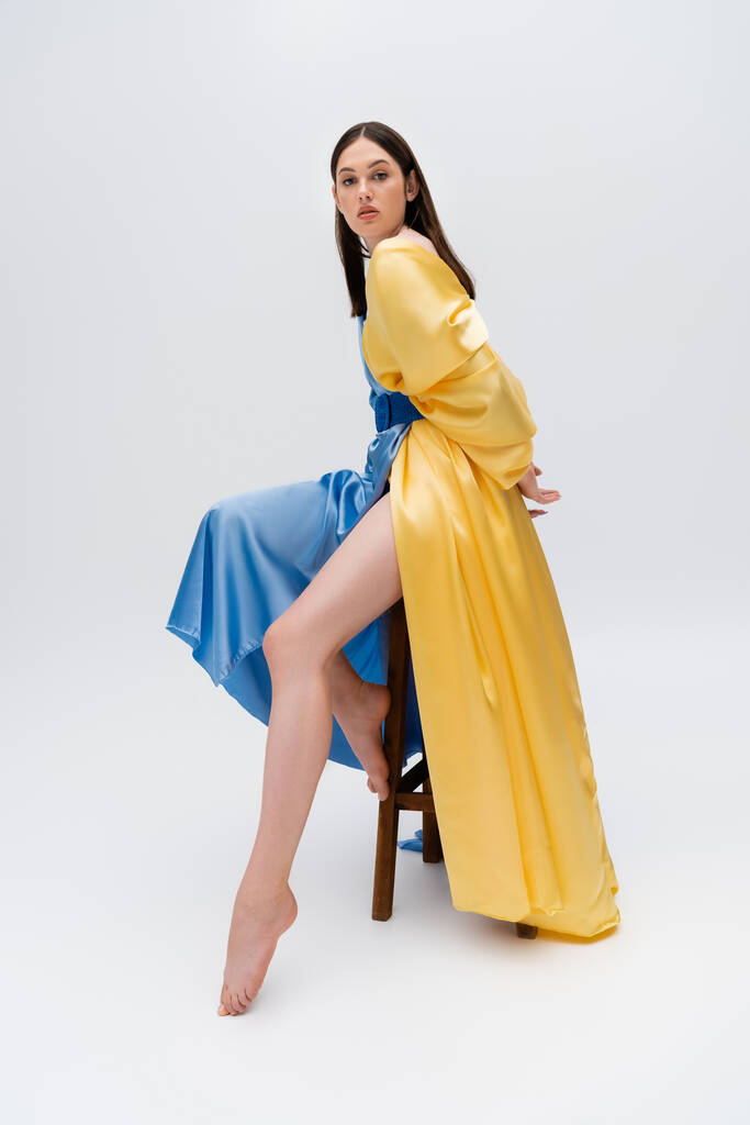 barefoot young ukrainian woman in blue and yellow dress posing while sitting on wooden chair on grey - Foto, afbeelding
