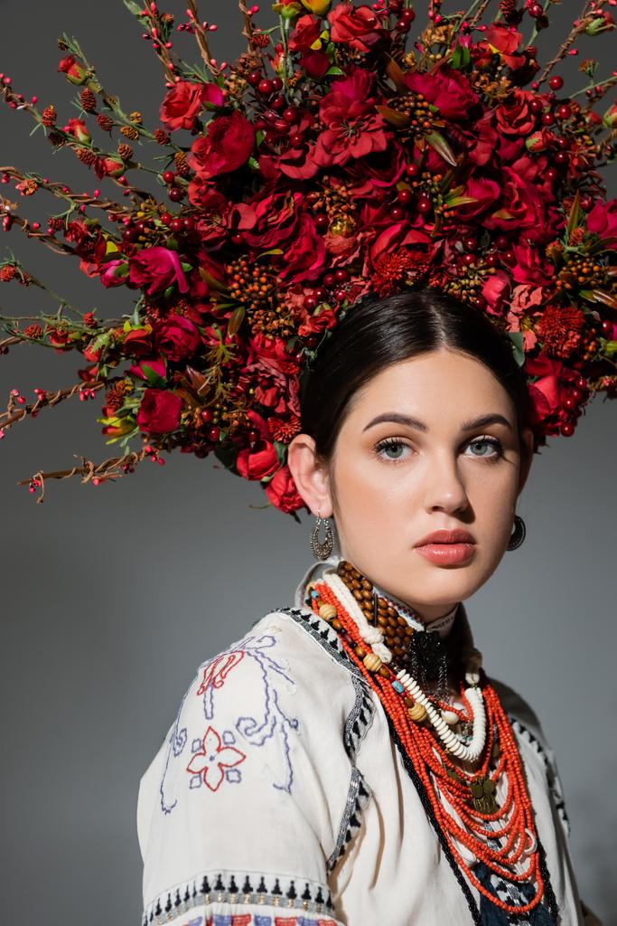portré csinos ukrán nő hagyományos ruházat és virágos vörös koszorú elszigetelt szürke - Fotó, kép